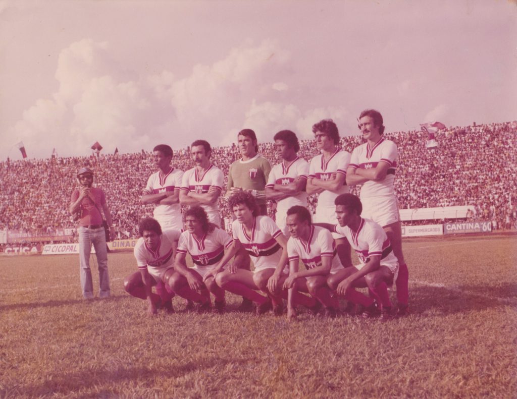 repórter de campo felicio papa em pé ao lado do time do botafogo de ribeirao preto do ano de 77, com destaque para sócrates agachado